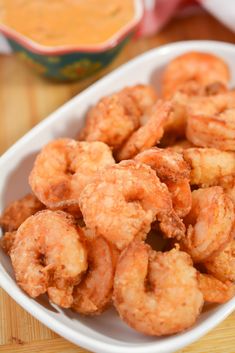 a white bowl filled with fried shrimp on top of a wooden table next to a container of dipping sauce