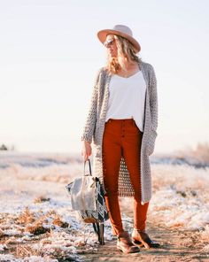 a woman standing in the snow with her hat on and holding a handbag next to her