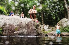 some kids are playing in the water near rocks and trees, while others look on