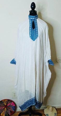 Ethiopian and Eritrean beautiful simple traditional  dress or cultural piece with blue and white patterns. It has a loose fit with decorative blue embroidery or trim at the neck, cuffs, and hem. Traditional White Kaftan For Spring, Traditional White Thobe With Patterns, Bohemian White Thobe For Spring, Spring White Tunic Thobe, White Folk Kaftan For Traditional Ceremonies, Traditional White Thobe For Summer, Traditional White Thobe For Vacation, Traditional White Thobe For Beach, Traditional White Summer Thobe