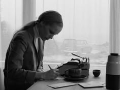 a woman sitting at a desk writing on a piece of paper with a typewriter in front of her