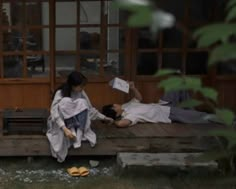 two people sitting on a wooden bench in front of a building and one person holding a paper