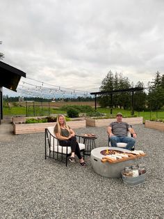 two people sitting in chairs around a fire pit with food on the table next to them