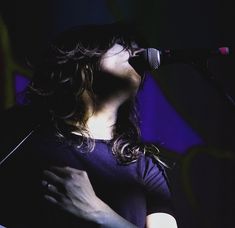 a woman singing into a microphone in front of a purple background with the light shining on her