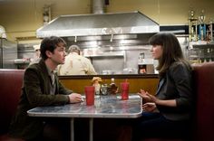 two people sitting at a table talking to each other in front of a restaurant kitchen