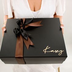 a woman is holding a large black box with a brown ribbon and name on it