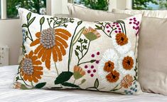 a white bed topped with two pillows covered in orange and green flowers next to windows