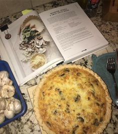 an open cookbook next to a pie on a counter with mushrooms and other food items