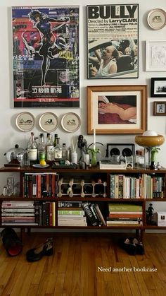 a book shelf filled with lots of books on top of a hard wood floor next to a wall covered in posters