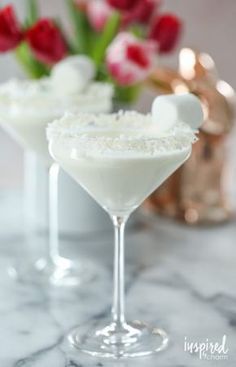 two martini glasses filled with white liquid on a marble counter top next to red tulips