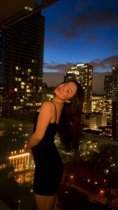 a woman standing on top of a building next to a tall building at night with city lights in the background