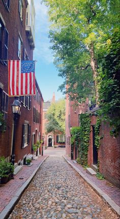 an empty cobblestone street with flags hanging from the buildings and trees on either side