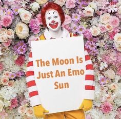 a clown holding a sign that says the moon is just an emo sun in front of flowers