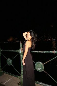 a woman in a long black dress standing on a bridge at night with her hands behind her head