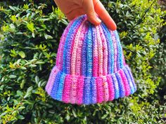 a hand is holding onto a multicolored knitted hat in front of bushes