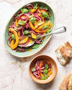 a bowl filled with sliced vegetables next to bread on a marble counter top and another bowl full of salad in the background