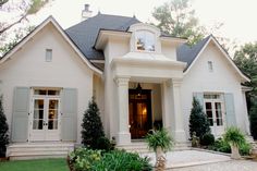 a white house with green shutters on the front door and windows, surrounded by greenery