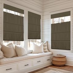 a window seat with pillows on it in front of two windows that have roman shades over them