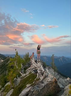 two people standing on top of a mountain with trees in the foreground and clouds in the background