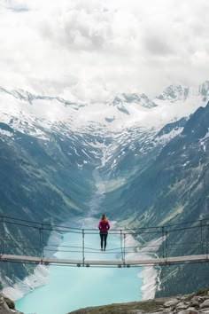 a woman standing on top of a bridge over a blue lake in the middle of mountains