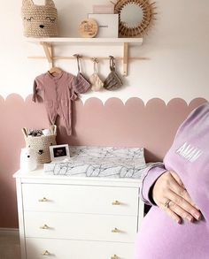 a pregnant woman standing in front of a dresser with baby clothes hanging on the wall