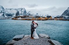 a bride and groom standing on rocks by the water in front of some houses with snow covered mountains behind them