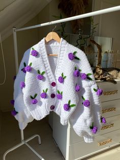 a white sweater with purple flowers is hanging on a clothes rack in front of a dresser