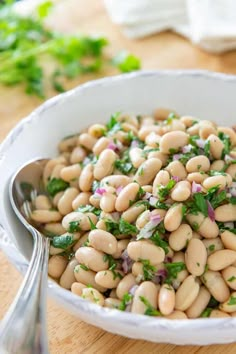 a white bowl filled with beans and greens on top of a wooden table next to a spoon