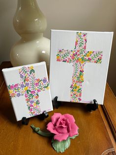 two cross paintings sitting on top of a wooden table next to a pink flower and a white vase