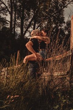 two people standing in tall grass next to each other near a wooden fence and trees