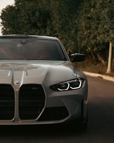 the front end of a silver sports car driving on a road with trees in the background