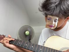 a young boy with his face painted like a clown holding an acoustic guitar and fan