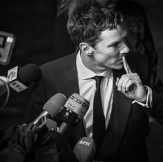 a man in a suit and tie holding a finger up to his mouth while surrounded by microphones