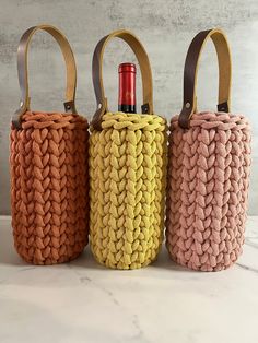 three knitted baskets with handles are lined up on a marble countertop, each holding a bottle of wine