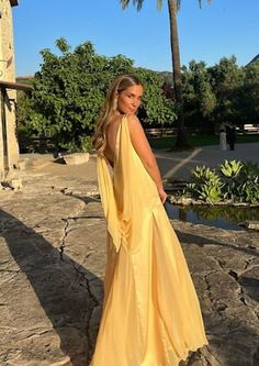 a woman in a long yellow dress standing on the rocks near some water and palm trees