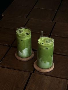 two glasses filled with green liquid sitting on top of a wooden table next to each other