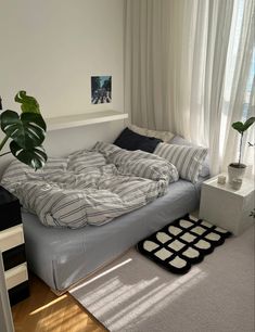 an unmade bed sitting in a bedroom next to a window with white curtains and a potted plant