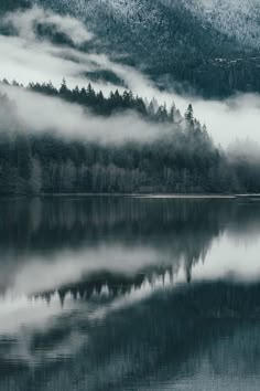 the mountains are covered in fog and low lying clouds above a lake with trees on both sides