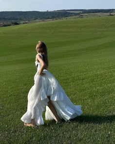 a woman in a long white dress walking across a lush green field with her back to the camera