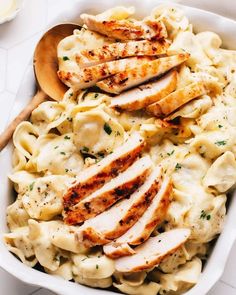 a white bowl filled with pasta and chicken breast slices on top of it next to a wooden spoon