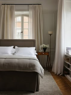 a bed with white sheets and pillows in a bedroom next to a window filled with curtains