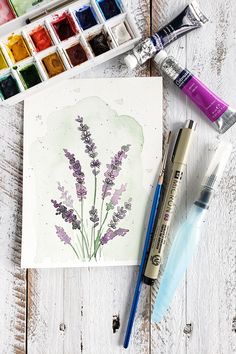 watercolor paints and markers are sitting on a table next to an art card with lavenders