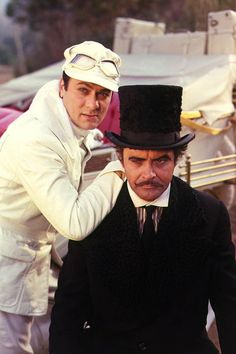 two men dressed in white and black, one wearing a top hat while the other is adjusting his tie