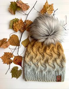 a knit hat with a pom - pom sits next to autumn leaves