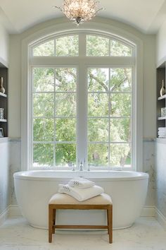 a white bath tub sitting under a large window next to a wooden footstool