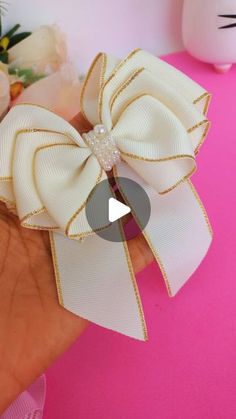 a hand holding a white and gold bow on it's left wrist with flowers in the background