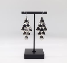 a pair of silver earrings hanging from a black stand on top of a white background