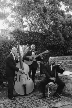 three men in suits and ties are playing instruments on the steps near some bushes, one man is sitting down