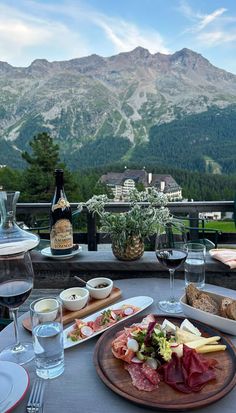 a table with food and wine on it in front of the mountains, overlooking a valley