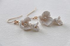 three white flowers are hanging from gold earwires on a white surface, with tiny pearls in the middle